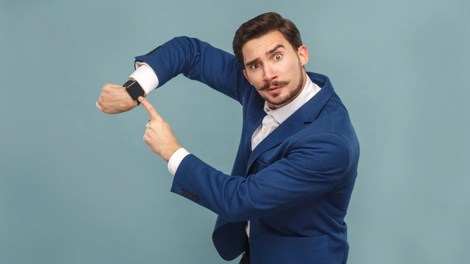 Un bărbat în costum de afaceri arată cu degetul spre ceasul de la încheietura mâinii.